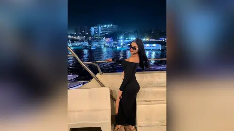 Stylish woman in a black off-the-shoulder dress posing on a boat at a night marina.