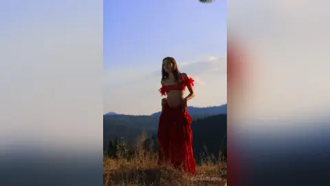 Elegant woman in a flowing red dress posing on a grassy hill under a clear blue sky at sunset.