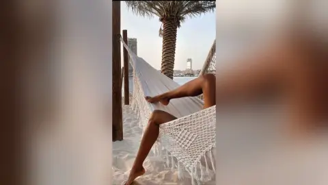 Relaxed woman in swimwear lounging in a white macrame hammock on a sunny tropical beach with palm trees.