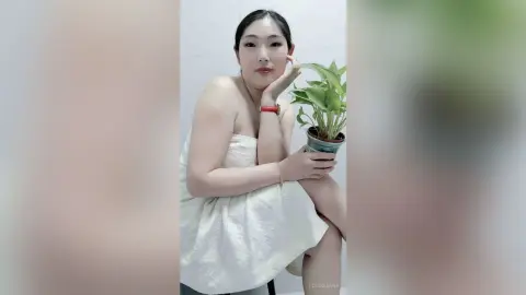 LouiseAnna poses naked with a white towel wrapped around her chest, resting her chin on her hand next to a small green plant.