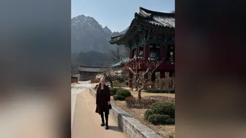 Woman in a black coat walking near a traditional Korean temple building with mountains in the background.