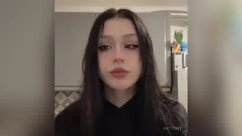 Young woman with long dark hair and winged eyeliner wearing a black top in a modern kitchen setting.