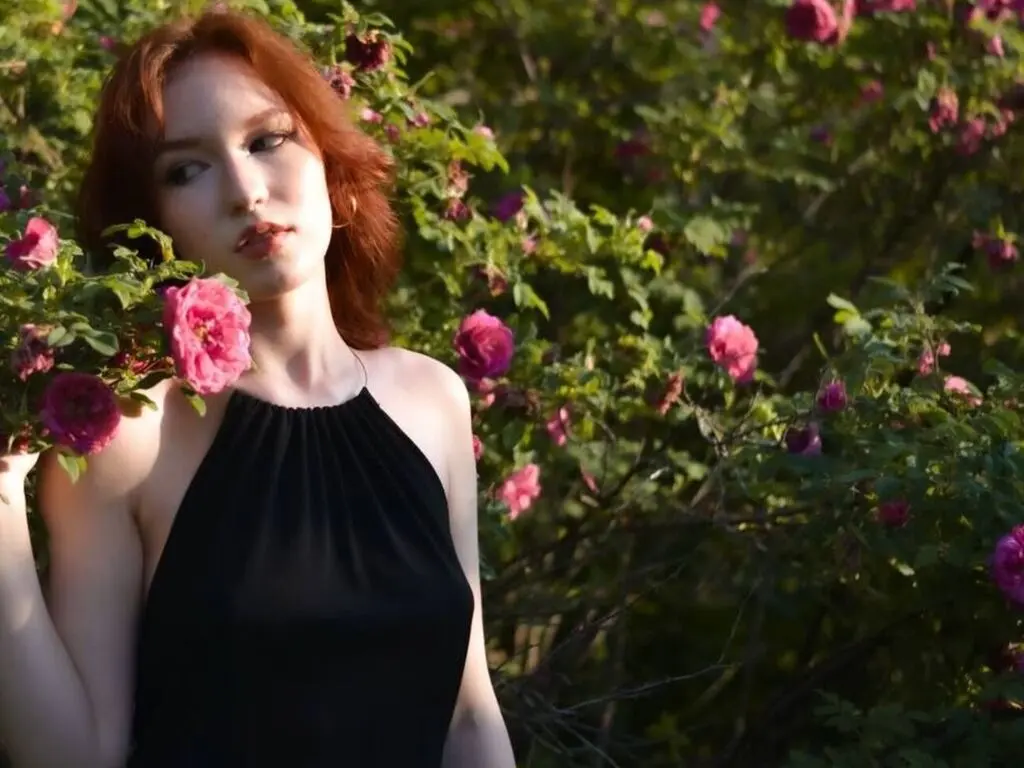 Redheaded woman in a black halter dress posing amidst blooming pink rose bushes in a dreamy garden setting.