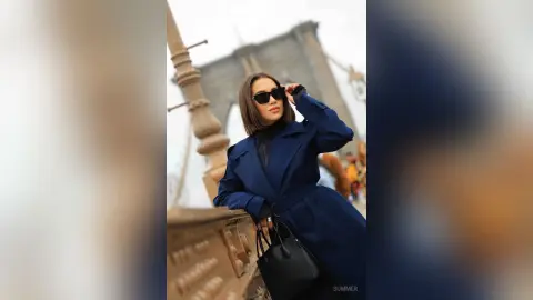 Stylish woman in a navy blue trench coat and sunglasses posing on a city bridge during a bright day.