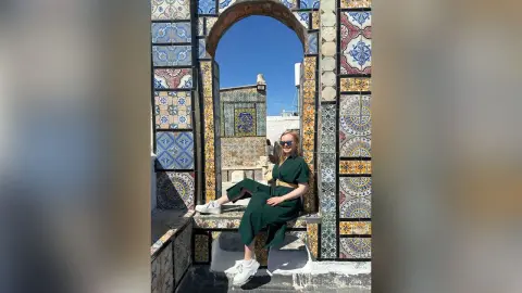 Blonde woman in a green dress sitting by colorful mosaic tiles under a bright blue sky in a Mediterranean courtyard.