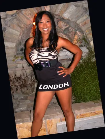 LondonSquirts smiles while wearing a black and white zebra-print mini dress with a black belt, standing in front of a stone archway.