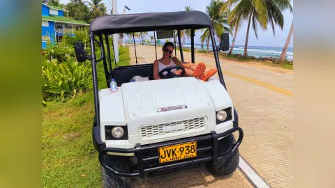 JessieLiu sits in a white golf cart on a paved road, wearing a white top and sunglasses with her legs resting on the front hood.