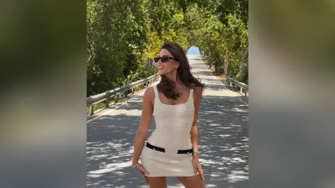 AnaMathers smiles while wearing a white tank-style mini dress and sunglasses on a sunlit road lined with green trees.