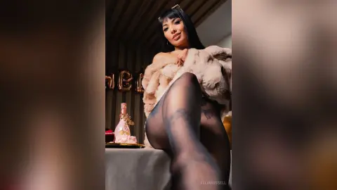 Glamorous woman in a beige faux fur coat and black tights sitting by a pink champagne bottle in a luxury setting.