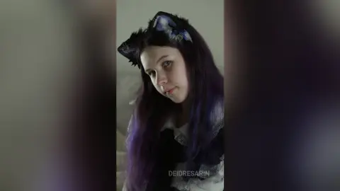 Young woman with purple hair wearing a black lace outfit and fuzzy cat ears in a soft indoor setting.
