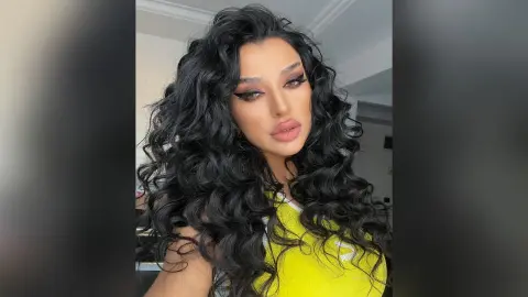 NicoleBella poses with long, wavy black hair wearing a bright yellow tank top.