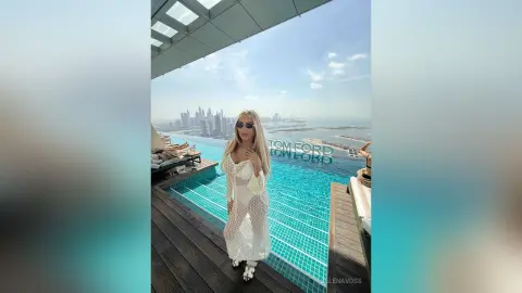 Blonde woman in a white lace maxi dress standing by a luxury rooftop infinity pool overlooking a city skyline.