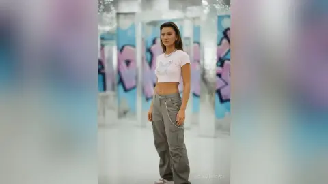 Young woman in a white butterfly crop top and grey cargo pants posing in a bright, modern studio.