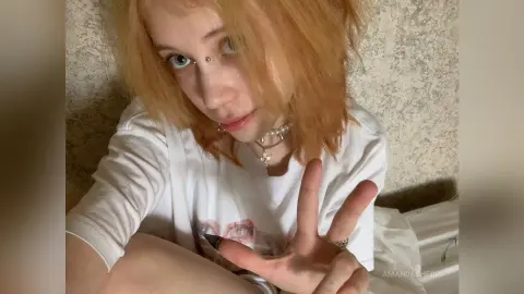 Blonde woman in a white graphic t-shirt making a peace sign in a cozy, casual indoor setting.