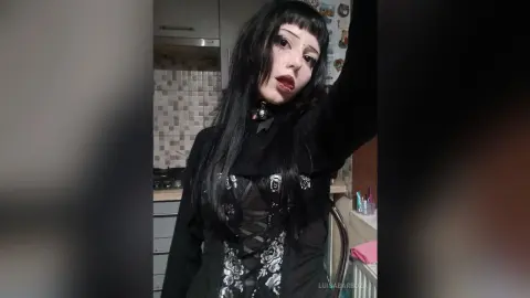 Gothic woman with long black hair wearing a black lace corset top in a dimly lit room.