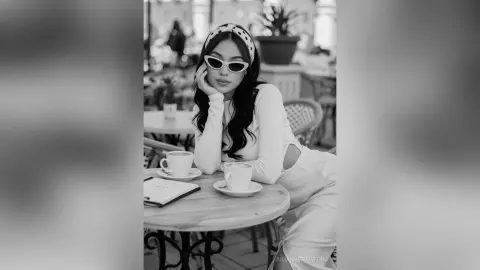 Stylish woman in white outfit and sunglasses sitting at a cafe table with coffee in a moody black and white portrait.