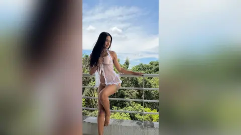 Young woman in a white lace negligee posing on a balcony overlooking lush green trees under a blue sky.