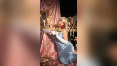 Elegant woman in a blue satin dress and white gloves holding a red rose in a dreamy, romantic studio setting.
