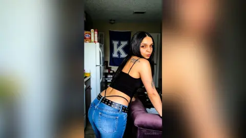 BiancaBell poses in a black tank top and blue jeans, showing off her back and tattoos in a kitchen setting.
