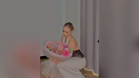 KrystalLaaz sits in a white dress holding a large bouquet of pink and white flowers.