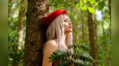 Dreamy portrait of a blonde woman in a red beret leaning against a tree in a lush green forest.
