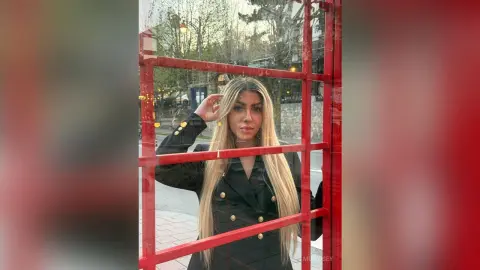 Blonde woman in a black blazer standing by a red window frame in an outdoor urban setting.