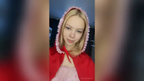 Blonde woman in a red hooded cape and pink dress smiling in a soft, whimsical indoor setting.
