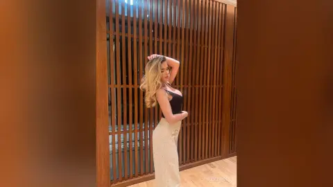 Blonde woman in a black tank top and white trousers posing against a modern wooden slatted wall.