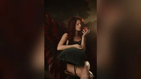 RubyAttwood poses with large red and black feathered wings, wearing a black lace bra and holding a red apple to her lips.