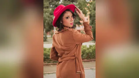 RebecaVillalobos wears a red hat and a tan belted coat while looking over her shoulder outdoors.