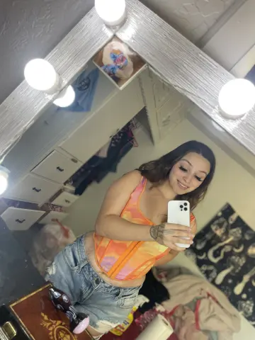 Princess1234 takes a mirror selfie wearing an orange and yellow tank top and denim shorts, showing her stomach and tattoos.