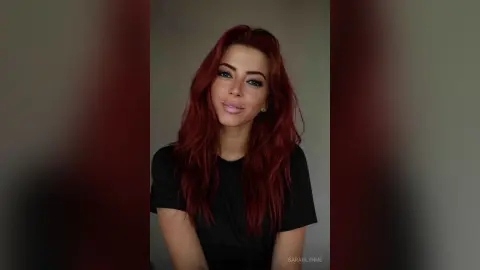 Smiling woman with long wavy red hair wearing a casual black t-shirt in a minimalist indoor setting.