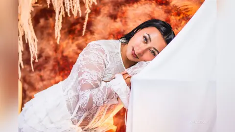 Elegant woman in a white lace long-sleeve dress leaning against a white fabric backdrop with a warm, dreamy mood.