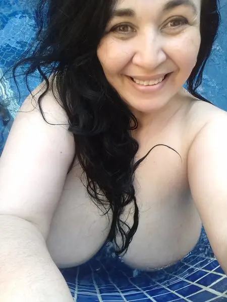 44DD_CICCIOLINA smiles while laying back, showing her long dark hair and cleavage in a dark top.
