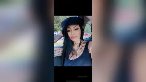 DeeaAnderson poses in a black tank top with long black hair and heavy eyeliner while sitting in a car seat.