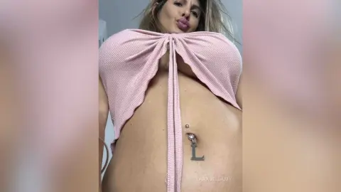 Young woman in a pink ribbed crop top showing her belly button piercing in a bright indoor setting.