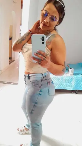 Woman in a white tank top and light blue jeans taking a mirror selfie in a bright bedroom.
