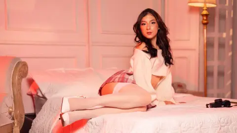 Young woman in a white sweater and plaid skirt relaxing on a bed in warm, moody pink lighting.