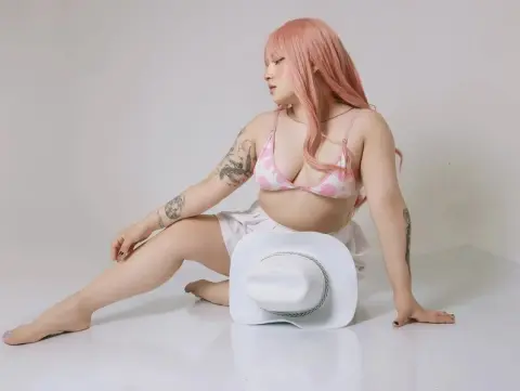 Pink-haired woman in a floral bikini and white shorts sitting in a bright studio with a white sun hat.