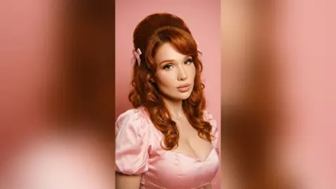 Redheaded woman in a vintage pink puff-sleeve dress with a retro hairstyle against a soft pink background.