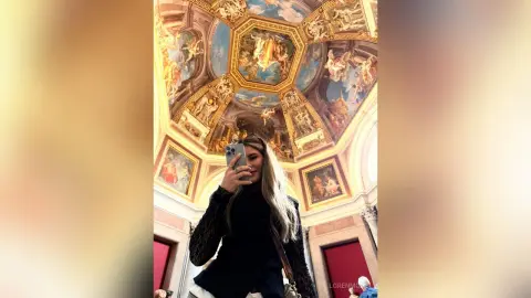 Blonde woman in a black lace top taking a selfie under a grand, ornate Renaissance fresco ceiling.