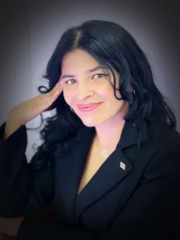 Smiling woman with long dark hair wearing a professional black blazer in a soft studio setting.