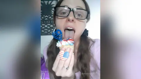 Young woman with glasses eating a patriotic cupcake with red, white, and blue frosting in a bright room.