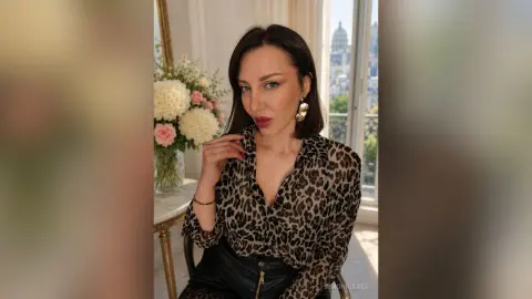 SimoniGrace poses in a leopard print blouse and black leather pants, looking at the camera in a bright room with flowers.