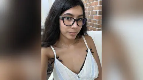 Young woman with glasses in a white camisole smiling in a bright indoor setting.