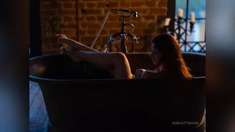 RubyAttwood relaxes in a dark bathtub with her legs raised, showing her bare legs and feet against a brick wall background.