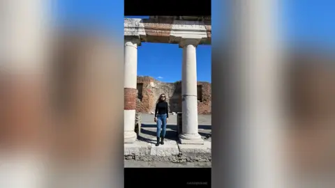 Woman in black long-sleeve shirt and blue jeans standing by ancient stone ruins under a bright blue sky.