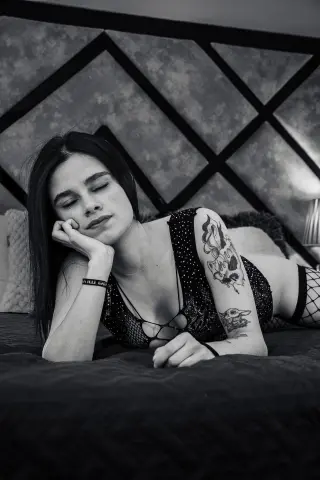 Tattooed woman in black polka dot lingerie relaxing on a bed in a moody, black and white bedroom setting.