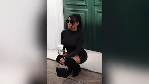 Stylish woman in a black long-sleeve dress and sunglasses holding a coffee cup by a dark green door.