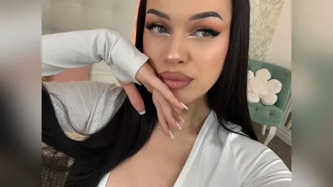 AriaVex poses with her hands near her face, wearing a white long-sleeved top and heavy eye makeup.
